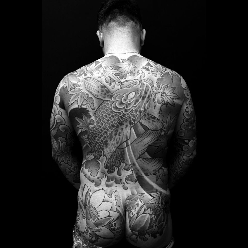 A man facing away from the camera, showcasing an extensive full-back and partial leg tattoo featuring a large koi fish surrounded by waves, lotus flowers, and traditional Japanese-style patterns, created in the style of a professional tattoo studio in Da Nang.