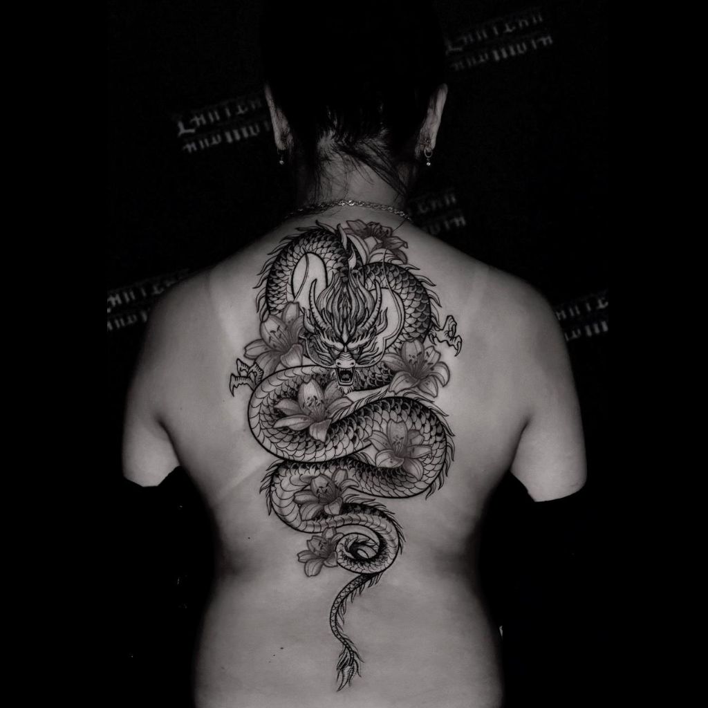 A woman with her back turned, showcasing an intricate full-back tattoo of a fierce, coiled dragon surrounded by flowers, photographed in a dark studio setting; artwork typical of a professional tattoo studio in Da Nang.